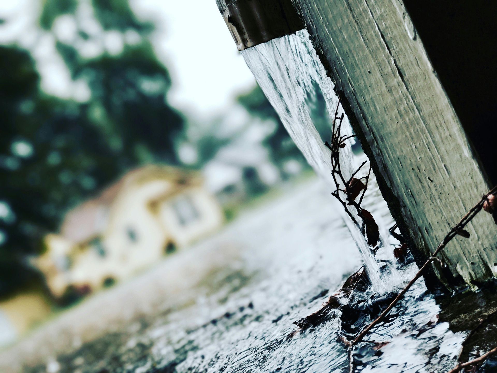 Rain runs down a wooden gutter. In the blurry background, a house and trees.